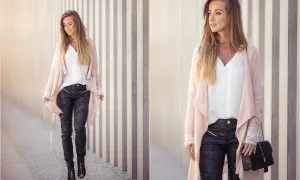 Elegancka kobieta w różowym kardiganie i czarnych spodniach woskowych z zamkami, spacerująca przy betonowej ścianie.