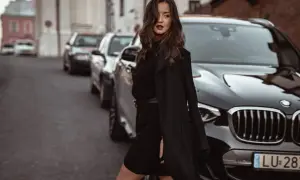 Elegancka kobieta w czarnej sukience swetrowej i płaszczu stoi obok luksusowego BMW na ulicy.