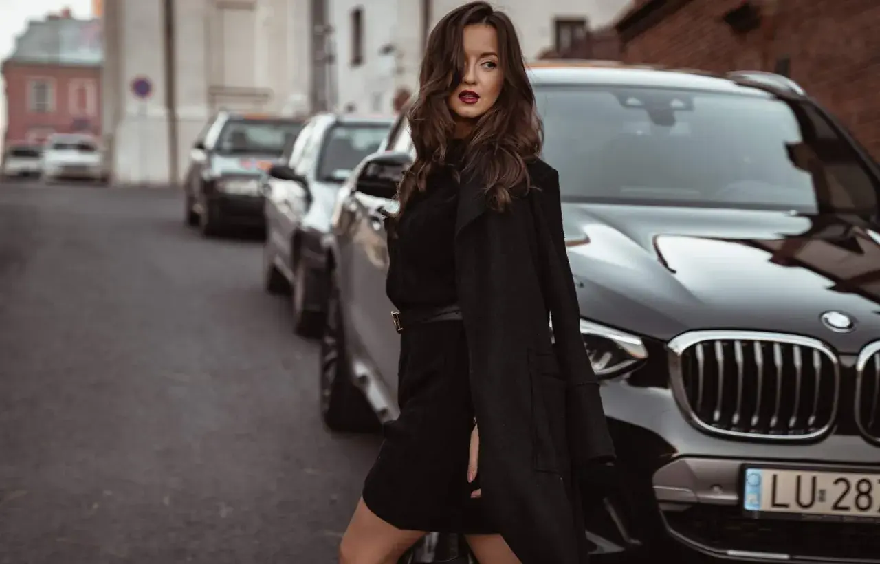 Elegancka kobieta w czarnej sukience swetrowej i płaszczu stoi obok luksusowego BMW na ulicy.